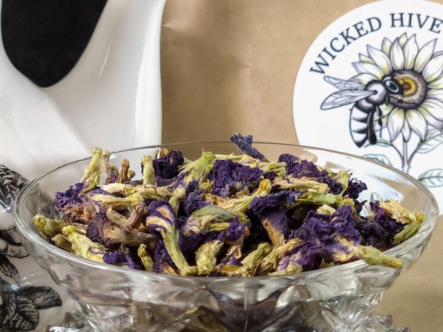 Dried butterfly pea flowers in a cut-glass bowl beside a Wicked Hive Garden kraft pouch.