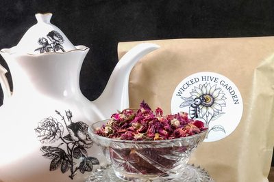 Dried rose buds in a cut-glass bowl beside a porcelain teapot and a Wicked Hive Garden pouch.